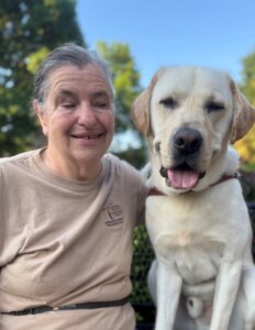 Handler Debra smiles as she sits next to yellow guide dog Gandalf who has a happy expression