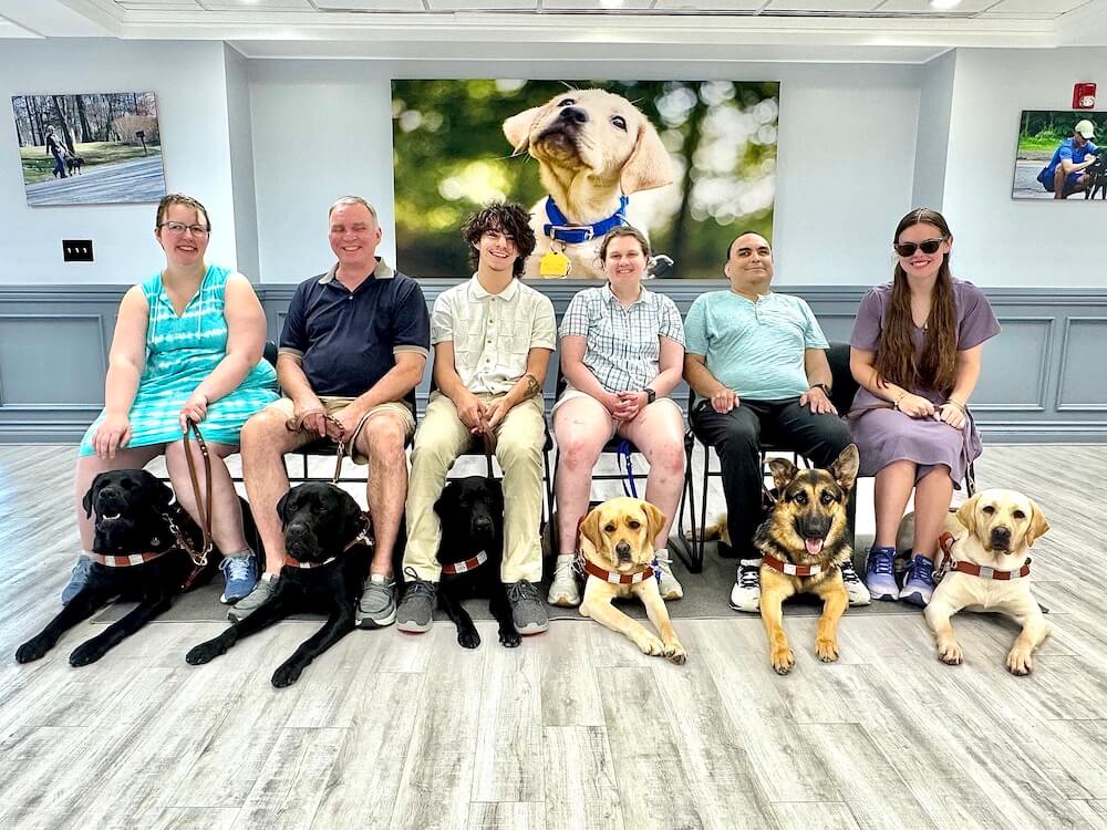 The six classmates of the July 7 2025 class sit in a line in chairs with their guide dogs in front of them.