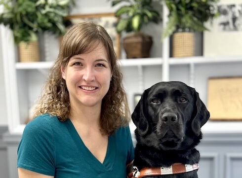 Rachel and black Lab guide dog Odyssey sit side by side in front of a backdrop of shelves with plants and other objects for their indoor team portrait