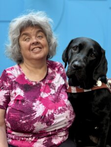 Sue Ellen and black Lab guide dog Chula sit in front of a blue background together for their team portrait