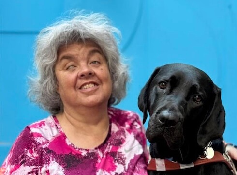 Sue Ellen and black Lab guide dog Chula sit in front of a blue background together for their team portrait