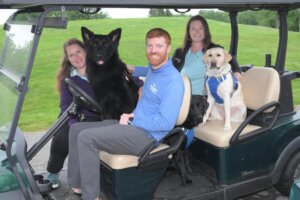 Instructors Dan Weesner, Allie Greenberg and Louise Thompson smile for the camera while sitting in a 2-row golfcart. A black German Shepherd sits on the front seat between Dan and Allie. A black lab sits on the floorboards with Louise, while a yellow lab sits next to her on the bench seat. 