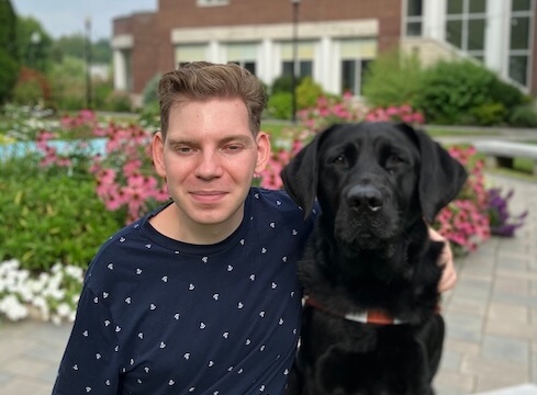 Ben and black Lab guide dog Viggo sit outdoors in a landscaped courtyard for their team portrait