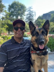 Dishon sits side by side with black and tan German Shepherd Rogue in an outdoor setting for their team portrait
