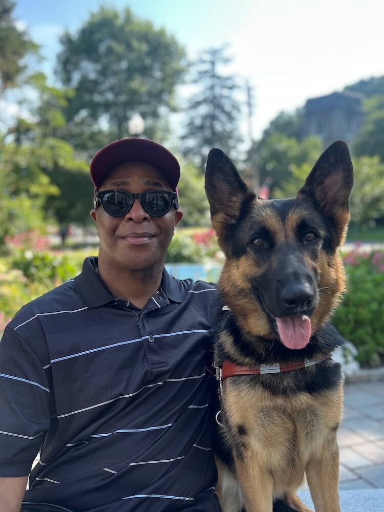 Dishon sits side by side with black and tan German Shepherd Rogue in an outdoor setting for their team portrait