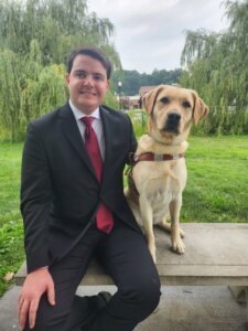 Robby and yellow Lab guide dog Fawn sit side by side on an outdoor bench in front of a grove of willows.