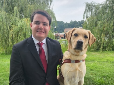 Robby and yellow Lab guide dog Fawn sit side by side in front of a grove of willows.