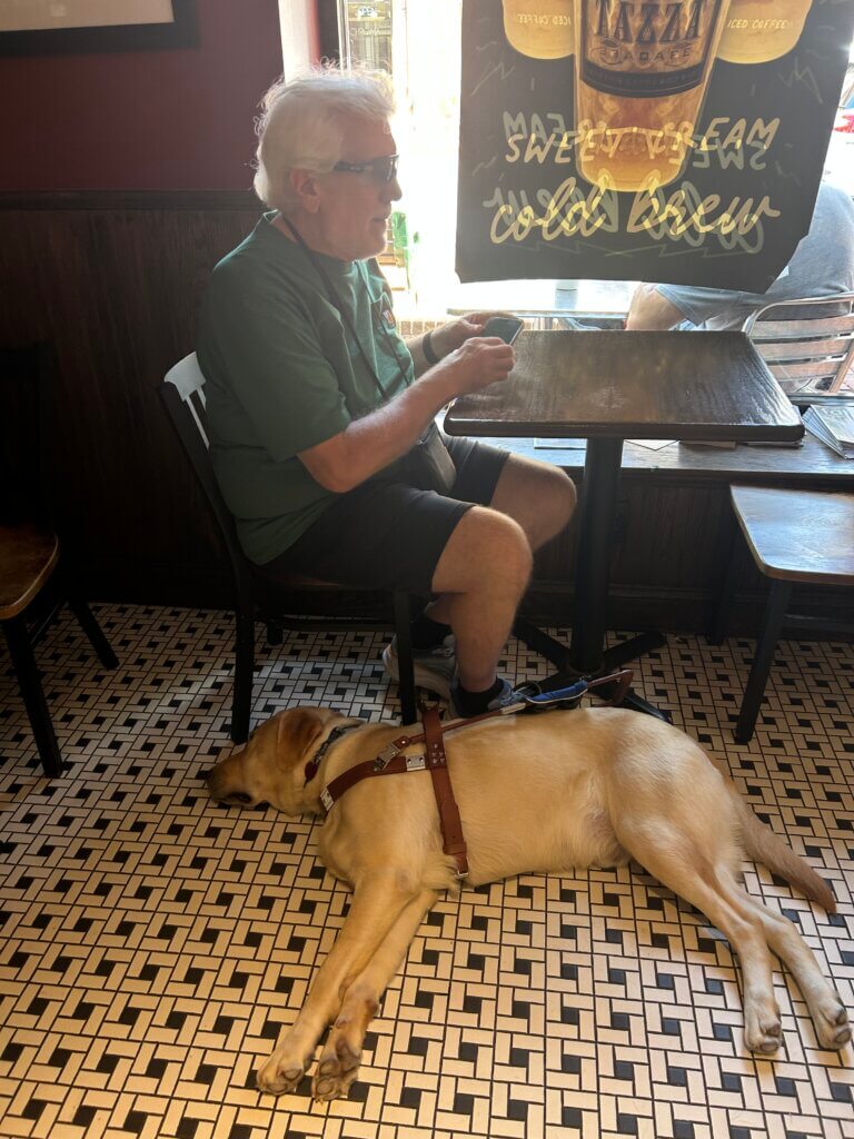 Terry works with his phone at a coffee shop table while guide dog Army stretches out on the floor