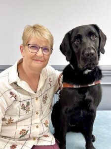 Connie sits beside black Lab guide dog Rhonda against a gray background for their team portrait