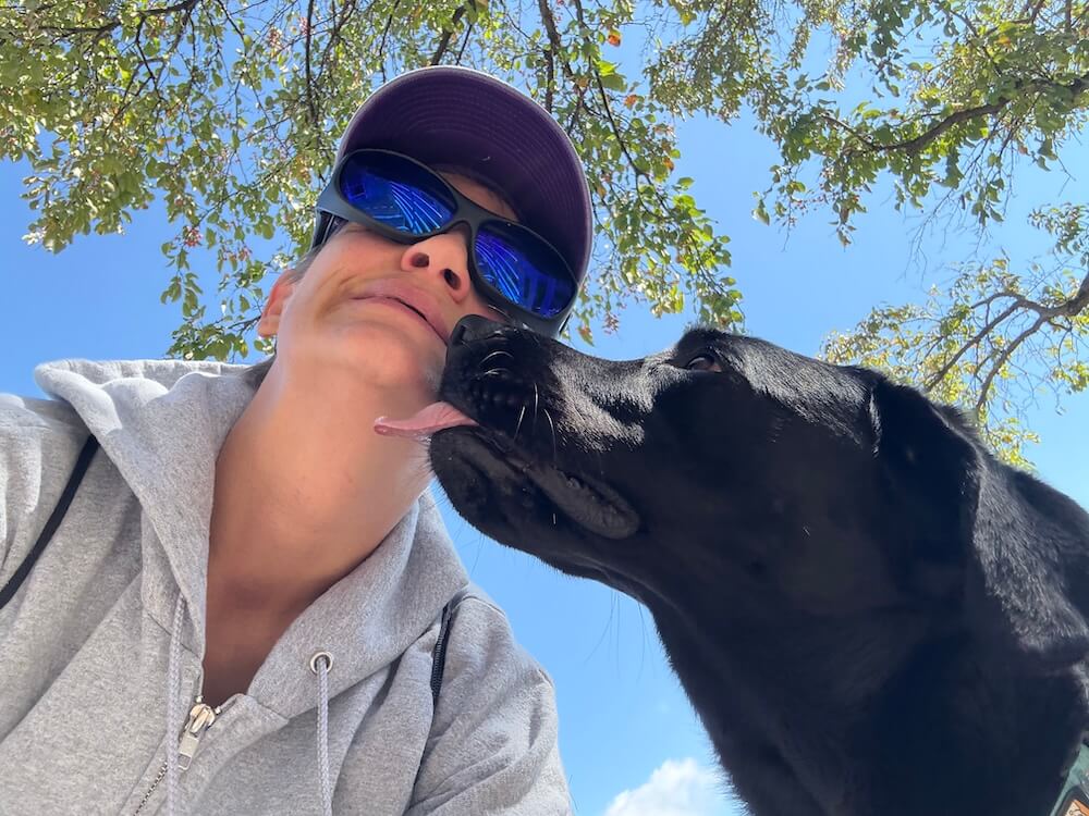 A graduate wearing blue sunglasses is kissed by her black Lab guide dog as the camera looks up to the tree limbs above them
