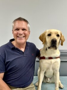 Joe sits beside yellow Lab guide dog Mitchell against a gray background for their team portrait