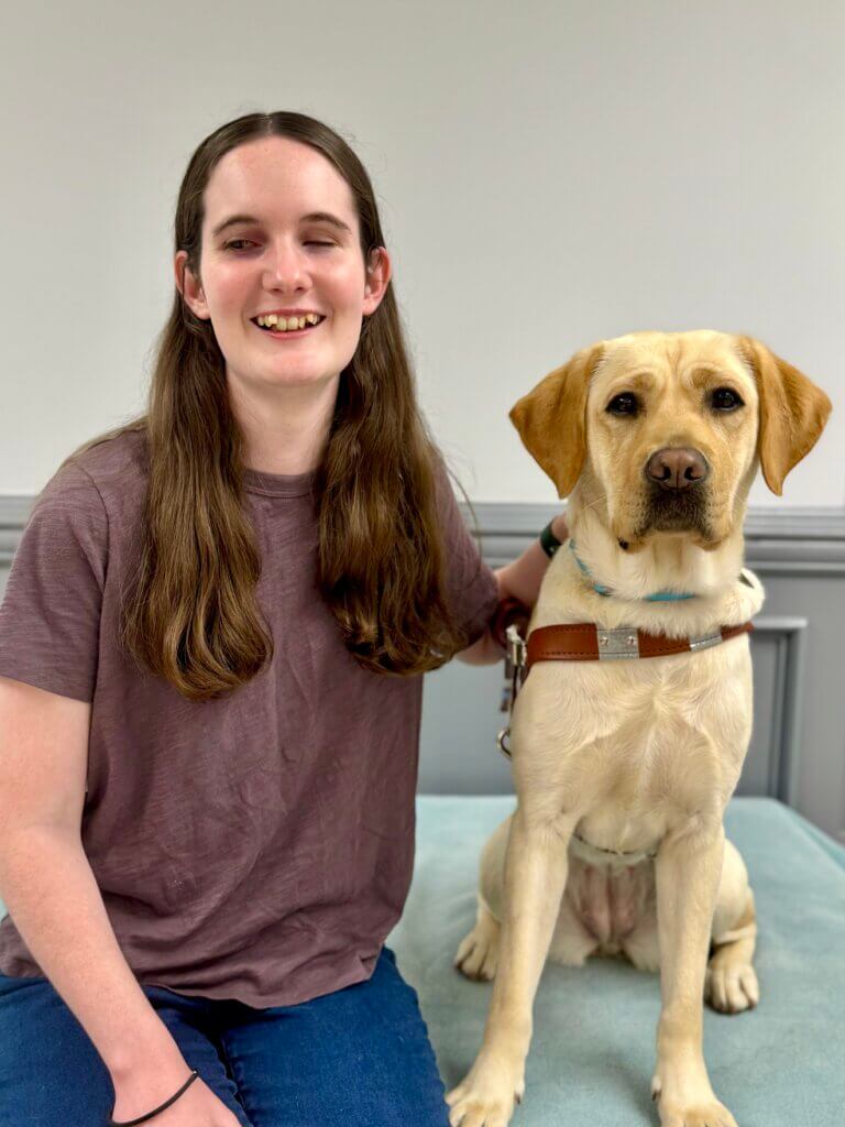Mia sits beside yellow Lab guide dog Melon, against a gray background, for their team portrait