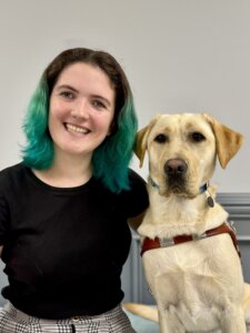 Abby sits beside yellow Lab guide dog Annie, against a gray background, for their team portrait