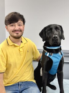 Christian sits beside black Lab guide dog Fudge against a gray background for their team portrait