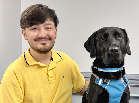 Christian sits beside black Lab guide dog Fudge against a gray background for their team portrait