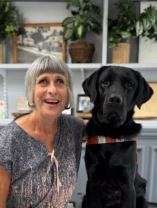 Deborah sits next to black Lab guide dog Everley against a backdrop of shelves for their team portrait