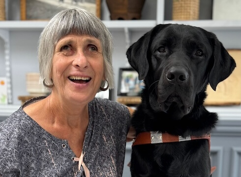 Deborah sits next to black Lab guide dog Everley against a backdrop of shelves for their team portrait