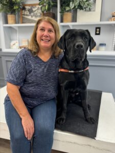 Kimberly sits next to her yellow Lab guide dog Quake in front of a bookshelf with plants and photos for their team portrait