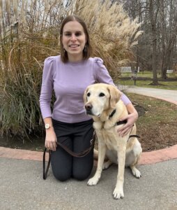 Melissa is looking directly at the camera with a nice, big smile, dressed in a pinkish/purplish sweater with gray pants and paw-print earrings, kneeling on the Guiding Eyes nature path in front of wispy, wheat-colored tall grasses. She has one arm around Aron, a yellow Lab who sits slightly facing towards the left, looking very sweet and gentle.