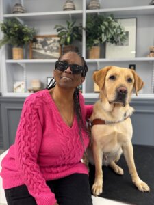 Pamela sits next to her yellow Lab guide dog Kingdom in front of a bookshelf with plants and photos for their team portrait