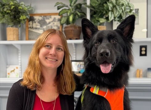 Shelby sits next to black German Shepherd guide dog Angel who wears a bright reflective harness, in front of a bookshelf with plants and photos for their team portrait