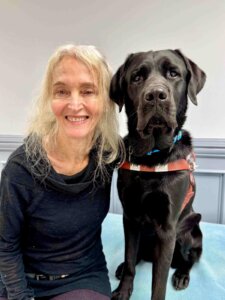 Hillary and black lab Essex are sitting and looking at the camera-Hillary is wearing a black sweater and has her left arm around Essex who is wearing a leather harness