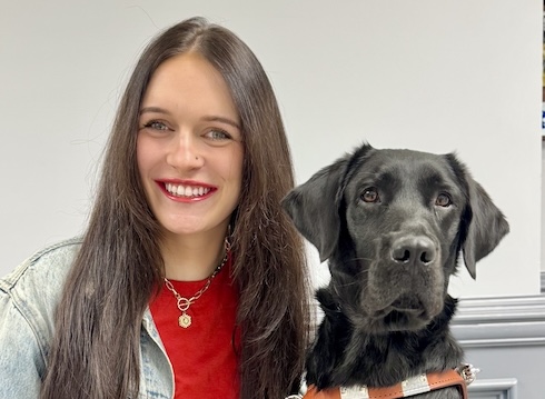 Olivia sits beside black Lab guide dog Omari against a gray background for their team portrait