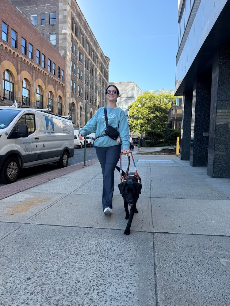 Olivia walks quickly and safely down a sidewalk on an urban street with guide Omari.