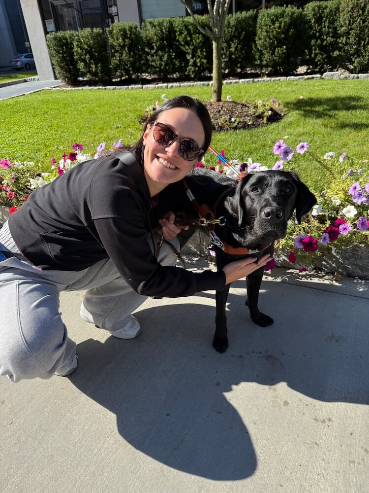 Olivia crouches down to hold black lab guide Omari in front of a garden of petunias