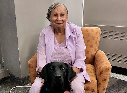 Sandra sits in a print upholstered arm chair with black lab Vega sitting in front of her.