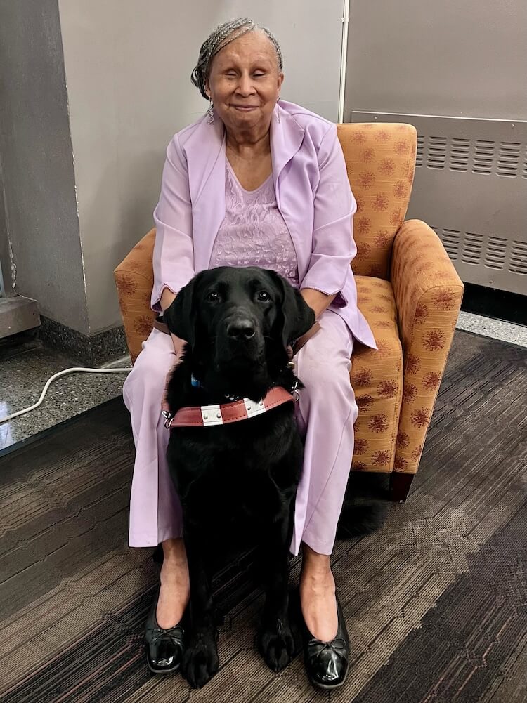 Sandra sits in a print upholstered arm chair with black lab Vega sitting in front of her.