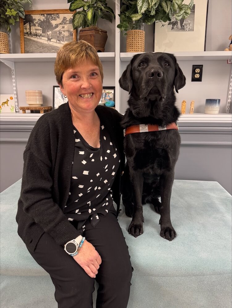 Tammy and black Lab guide dog Varsity sit in front of shelves with plants and pottery items for their team portrait