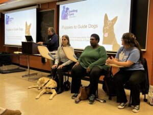 Cindy Tait Regional Puppy Instructor stands at the podium to begin the presentation while Melissa with guide dog Aron-Meka and Nikki sit in the front