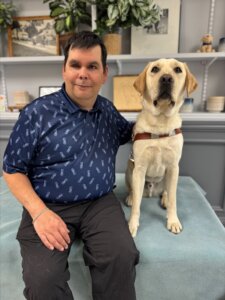 Adam and yellow Lab guide dog Beau sit in front of shelves with plants and framed items for their team portrait