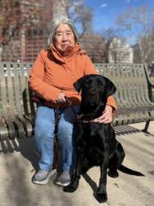 Helen sits on a metal park bench with her arm draped around black lab guide dog Quinoa at her feet for their team portrait