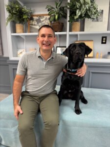 Jake and black Lab guide dog Fianna sit in front a bookcase with plants & various items for their team portrait