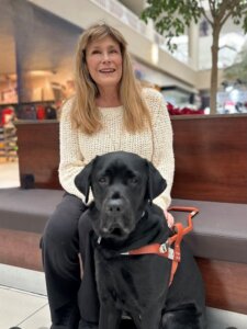 Cheryl and black Lab guide dog Helix sit on a bench in a mall for team portrait