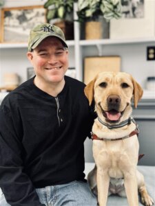 Dan and yellow Lab guide dog Lucia sit in front of shelves with various items for their team portrait