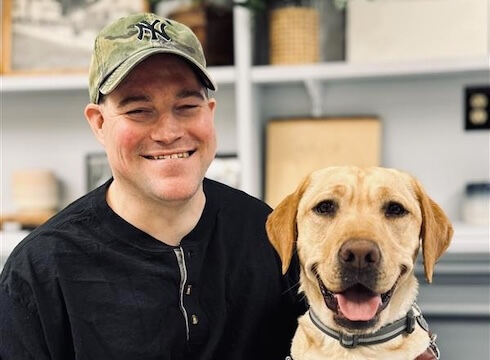 Dan and yellow Lab guide dog Lucia sit in front of shelves with various items for their team portrait