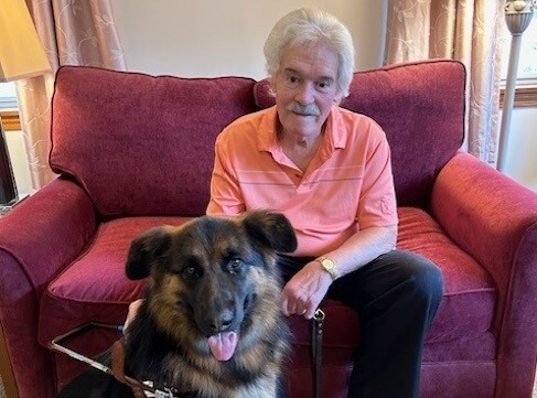Marc sits in an oversized red chair as German Shepherd guide dog Idril sits happily at his feet