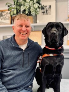 Mark and black Lab guide dog Denzel sit in front of shelves with plants and pottery items for their team portrait
