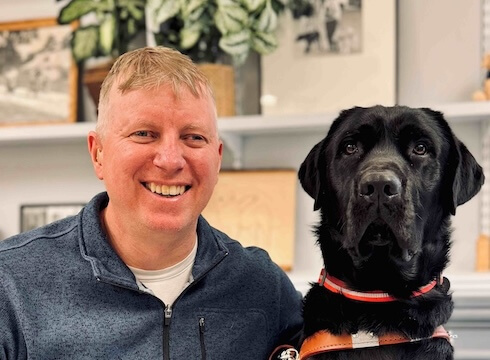 Mark and black Lab guide dog Denzel sit in front of shelves with plants and pottery items for their team portrait