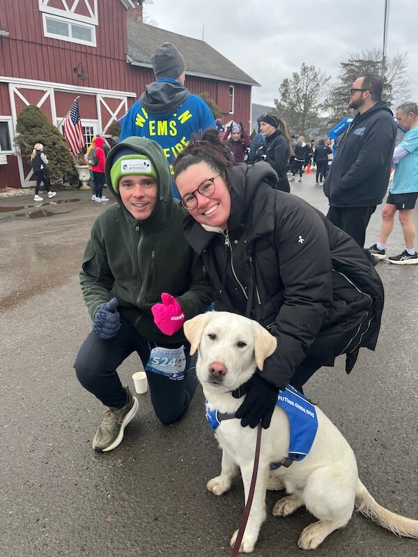Pup Polar in a Future Guide Dog vest poses with two attendees with his usual calm gaze at the camera