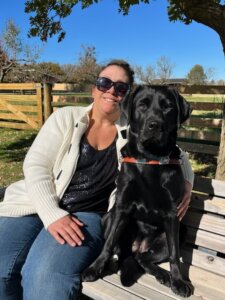 Rebecca and black Lab guide dog Gia sit together on a wooden bench in front of wood fencing for their team portrait
