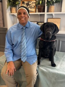 Amir and black Lab guide dog Irving sit for their team portrait in front of shelves filled with various items 