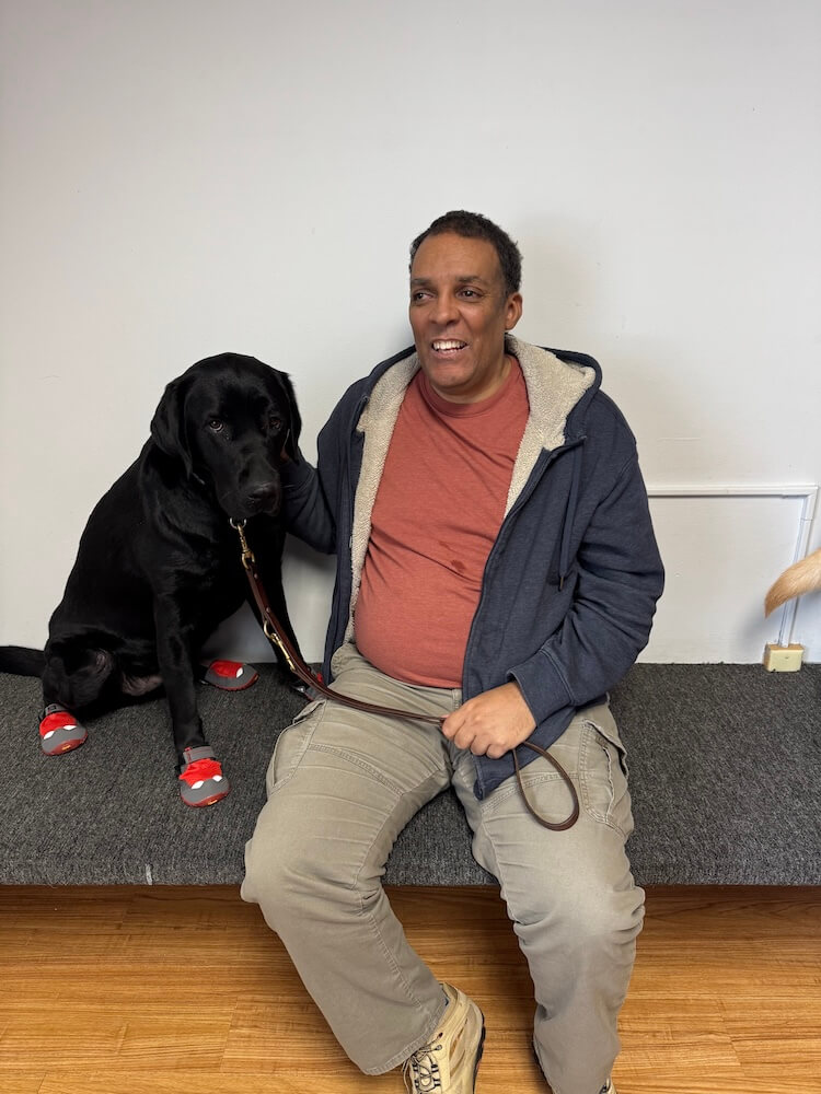 Black lab guide dog Irving sits next to Amir on the grooming bench wearing booties
