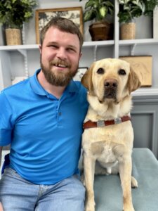 Austin and yellow Lab guide dog Jimbo sit in front of shelves filled with various items for their team portrait