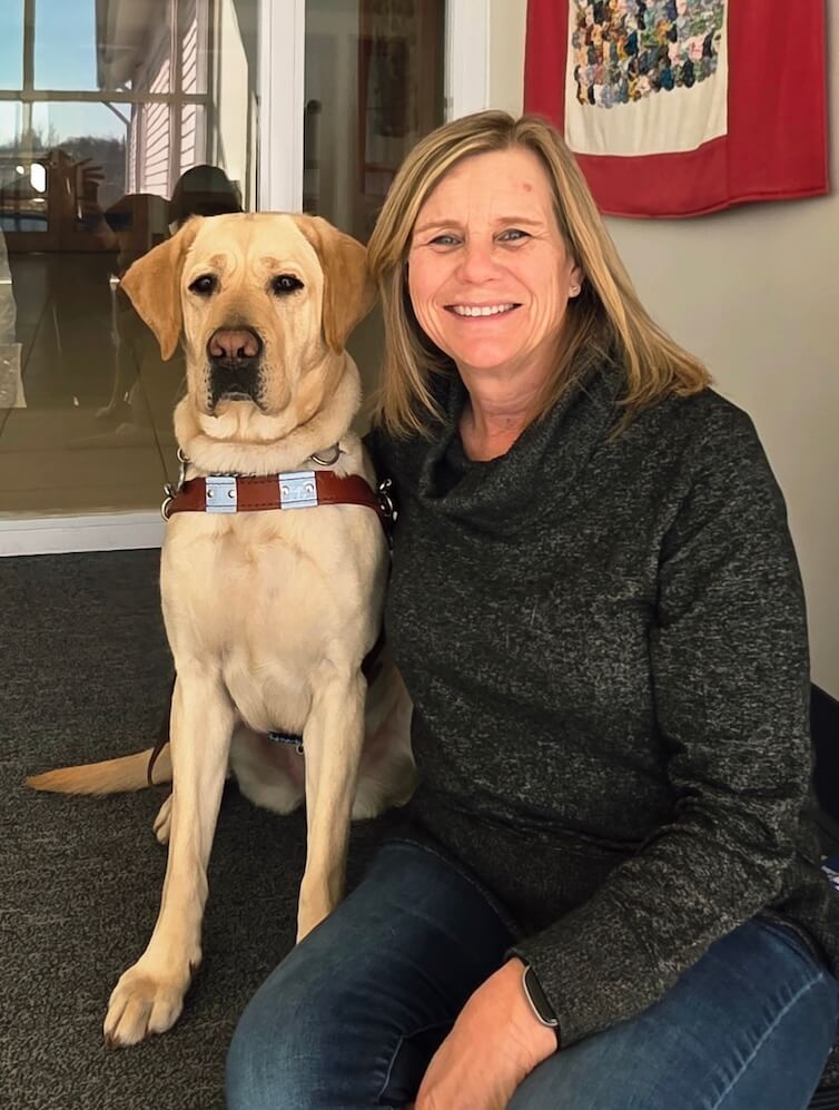 Carol and yellow guide dog Mia sit on a carpeted step in front of a wall of windows for their team portrait