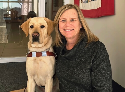 Carol and yellow guide dog Mia sit on a carpeted step in front of a wall of windows for their team portrait