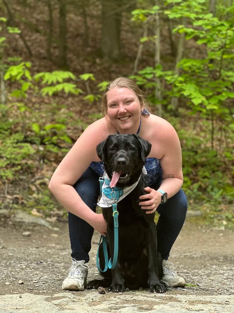 Irving sits in a wooded path with his puppy raiser Char